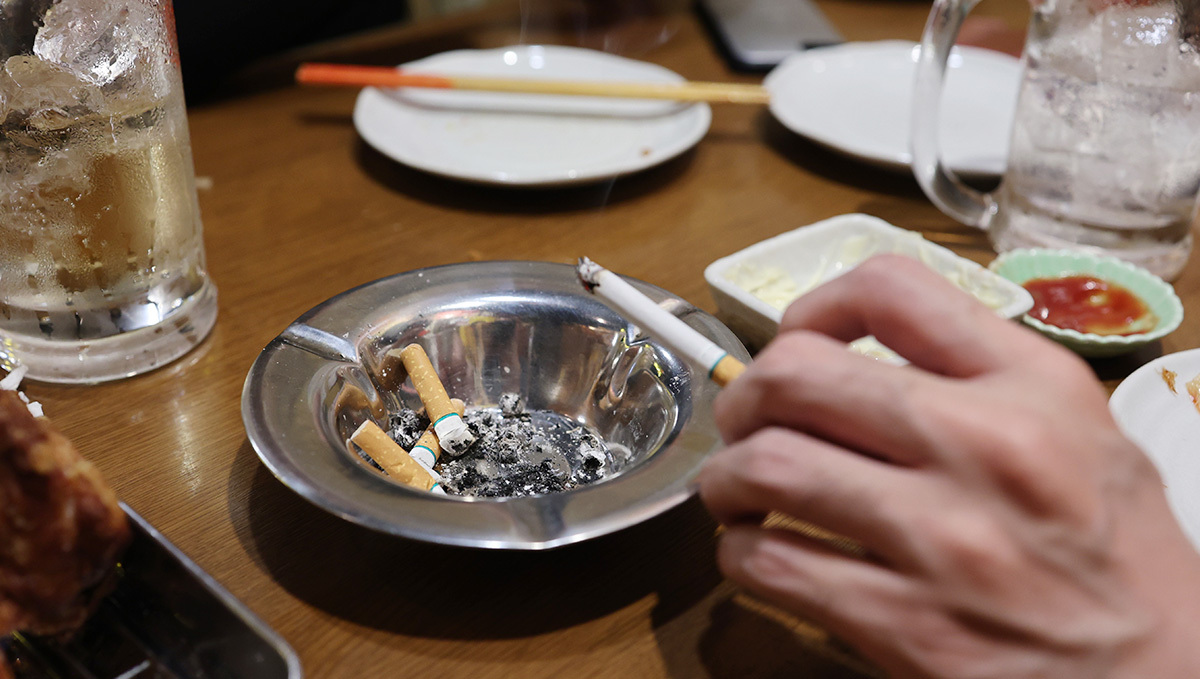 Weed Taxes Bring In 3x More Than Alcohol + Tobacco in Arizona: A closeup of a tabletop with an alcoholic drink and an ashtray; a hand is seen shaking ashes from a burning cigarette