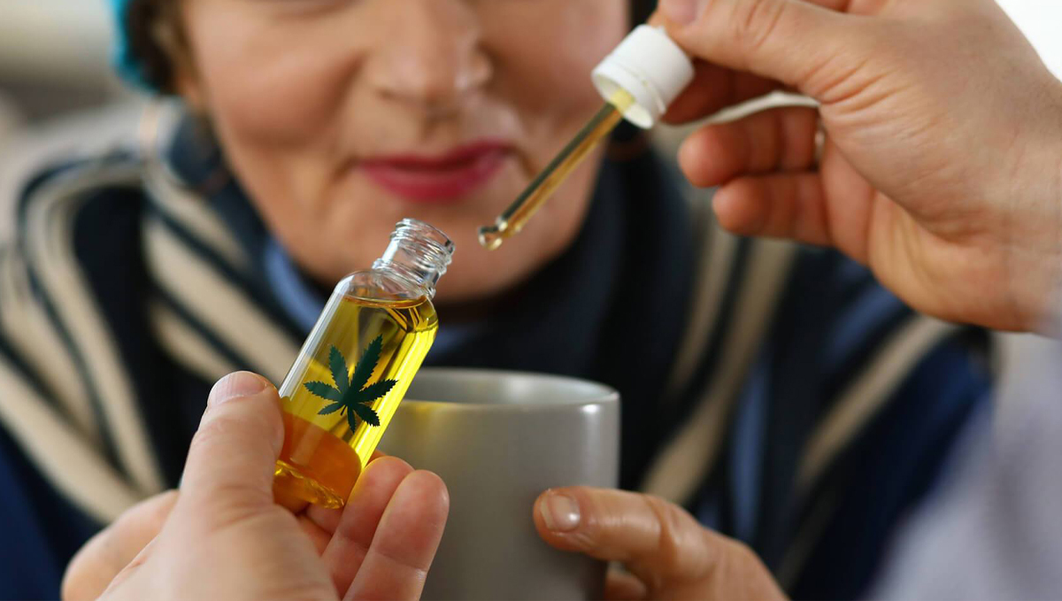Cannabis Is Becoming More Popular Among the Elderly: A caregiver adding cannabis oil in a cup that an elderly woman holds Cannabis Is Becoming More Popular Among the Elderly: A caregiver adding cannabis oil in a cup that an elderly woman holds